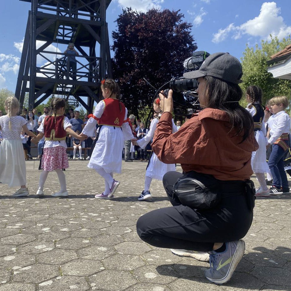 The image shows a woman filming a cultural presentation where children are dancing and spinning.